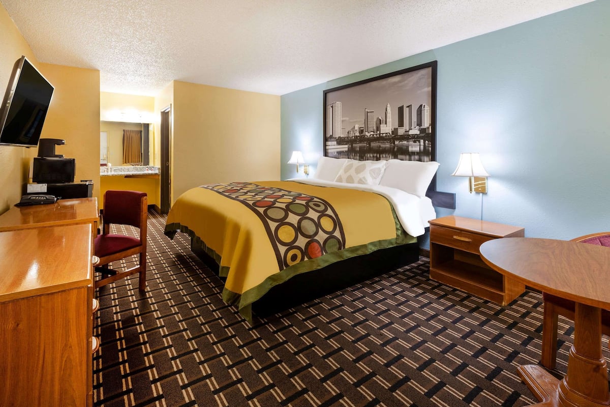 A hotel room with a bed, a desk, a chair, a TV, and a cityscape painting on the wall.