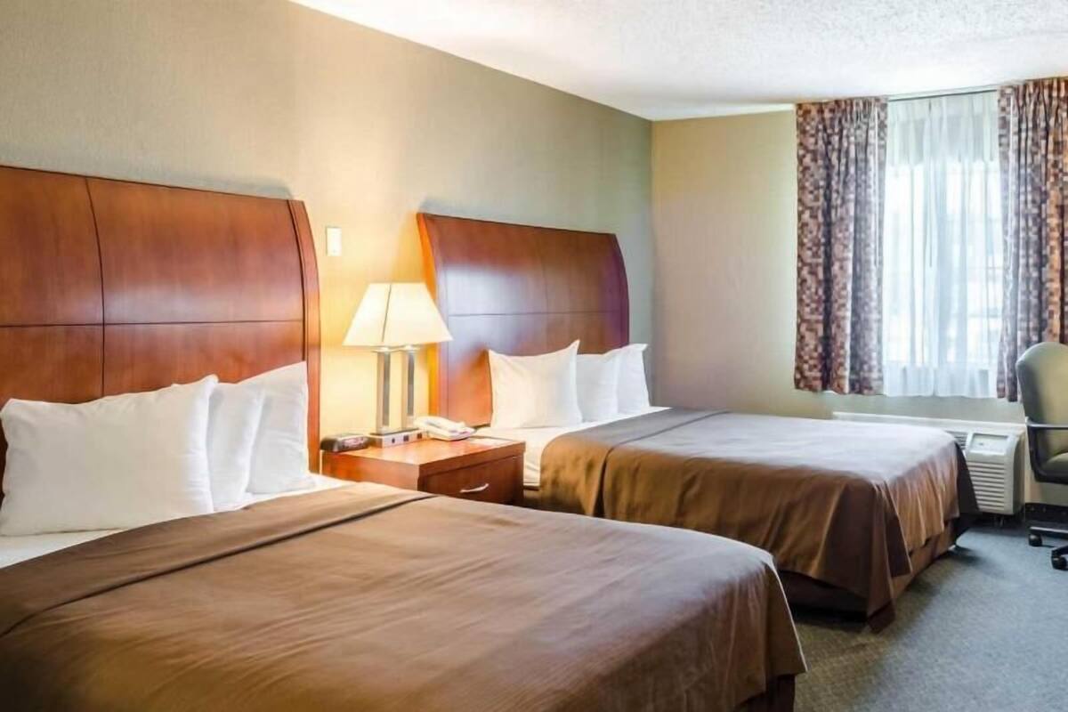 A hotel room with two beds, a nightstand with a lamp, a chair, and a window with curtains.