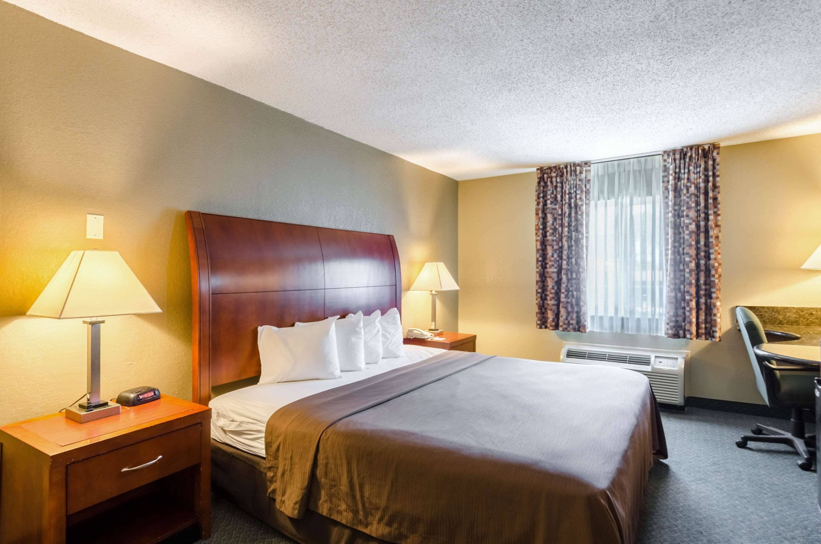 A hotel room with a large bed, two bedside tables with lamps, a desk with a chair, and a window with curtains.
