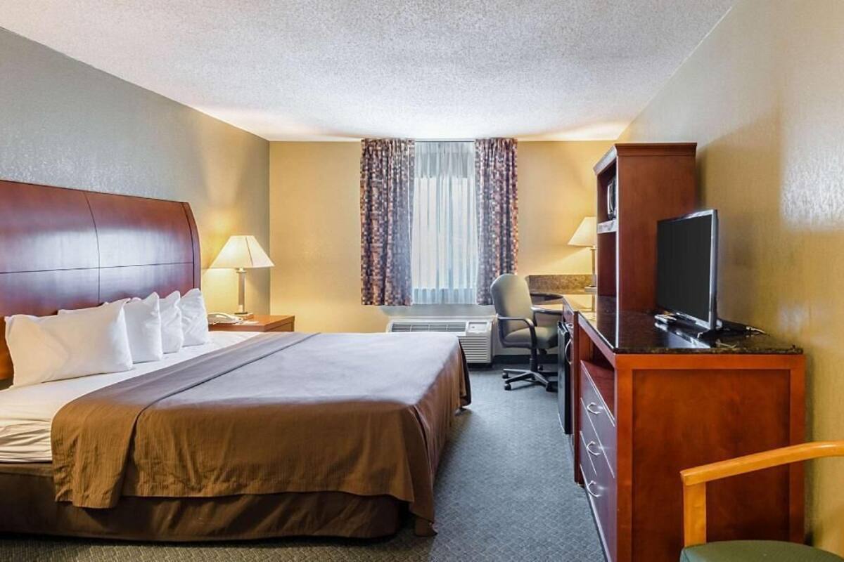 A hotel room with a bed, desk, chair, television, and air conditioning unit.