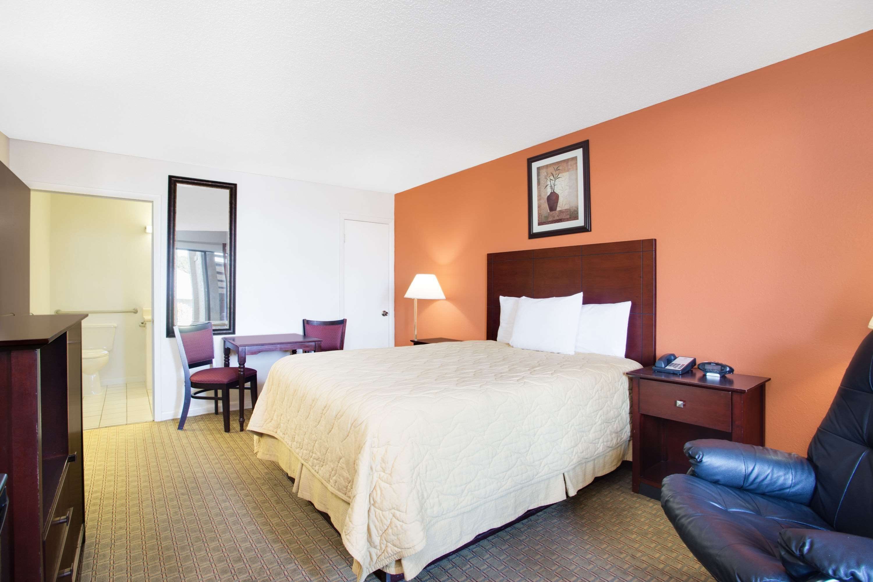 A hotel room with a bed, a desk, a chair, a nightstand, and a large mirror.