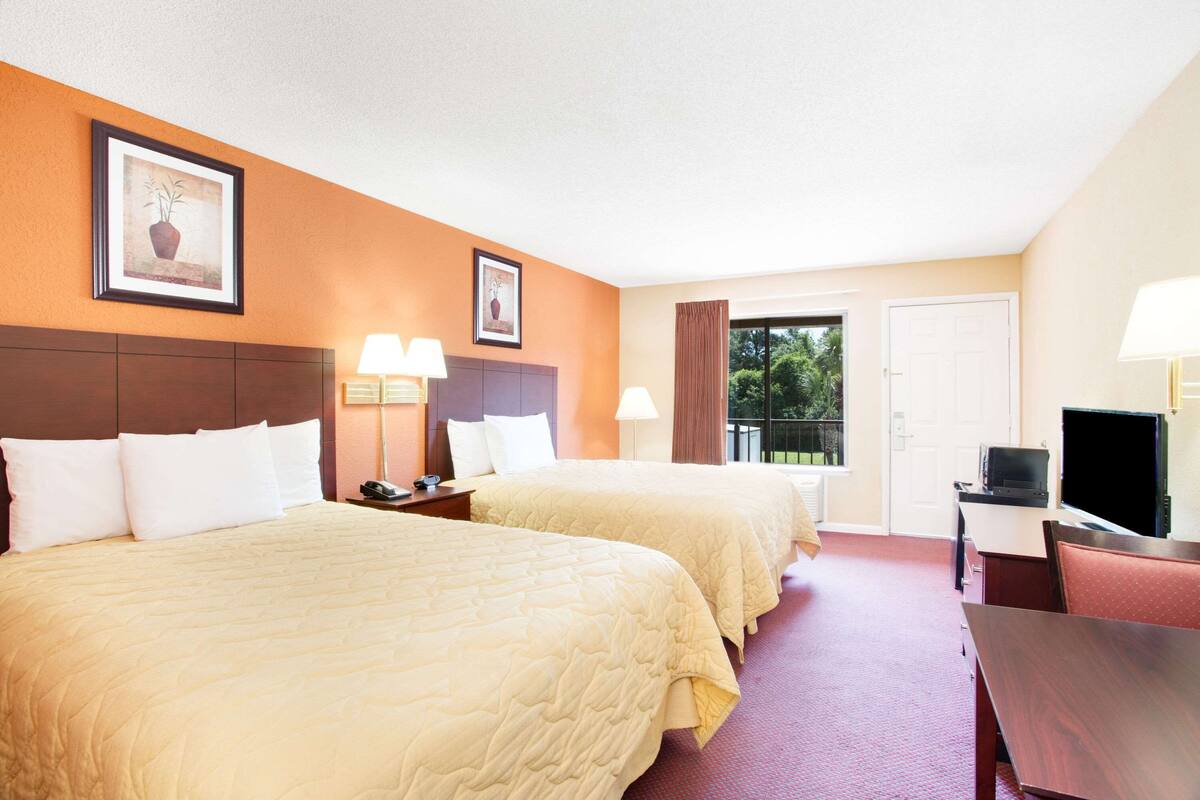 A hotel room with two beds, a desk, a TV, and a window with a view of greenery.