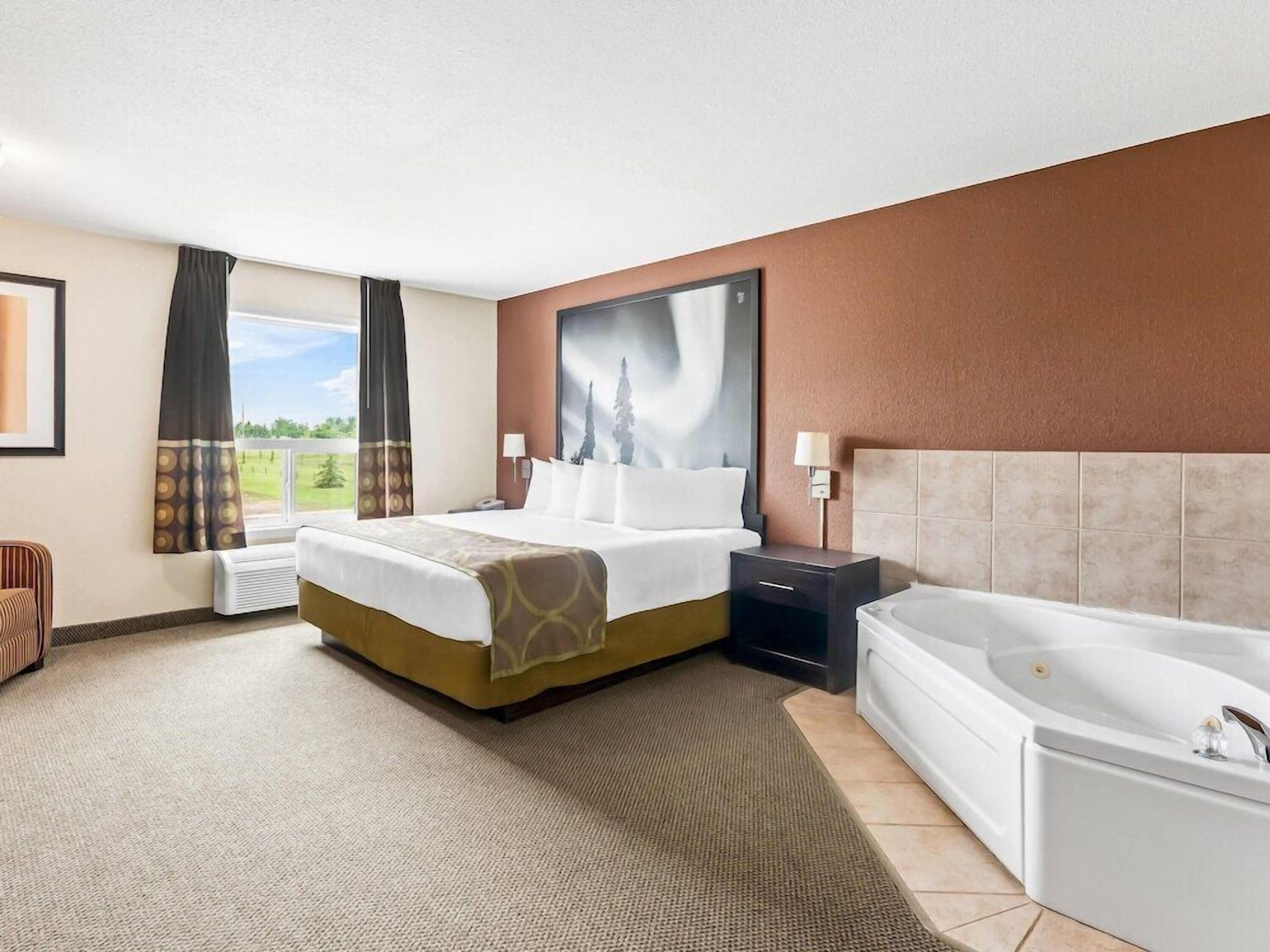 A hotel room with a bed, a large window, a painting, a nightstand, and a bathtub.