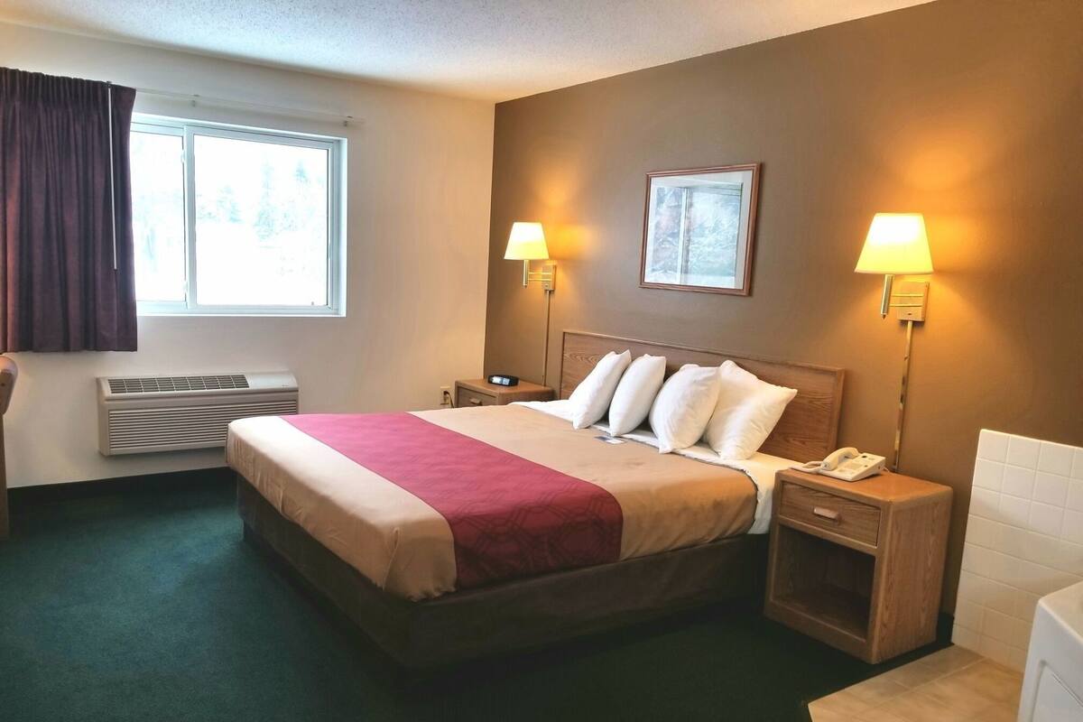 A hotel room with a bed, two bedside lamps, a nightstand with a phone, a window with curtains, and an air conditioning unit.