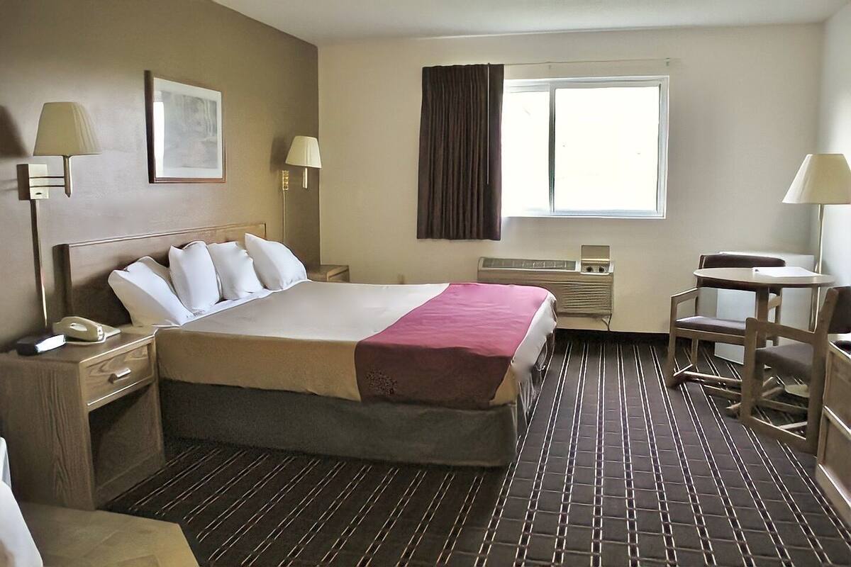 A hotel room with a bed, bedside tables, a desk, a chair, a window, and a wall-mounted air conditioner.