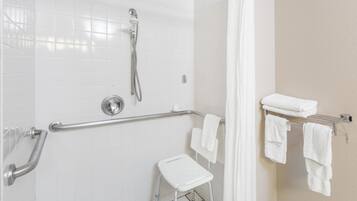 A bathroom with white tiled walls, a shower with a handheld showerhead, a grab bar, a white step stool, and a towel rack with folded towels.