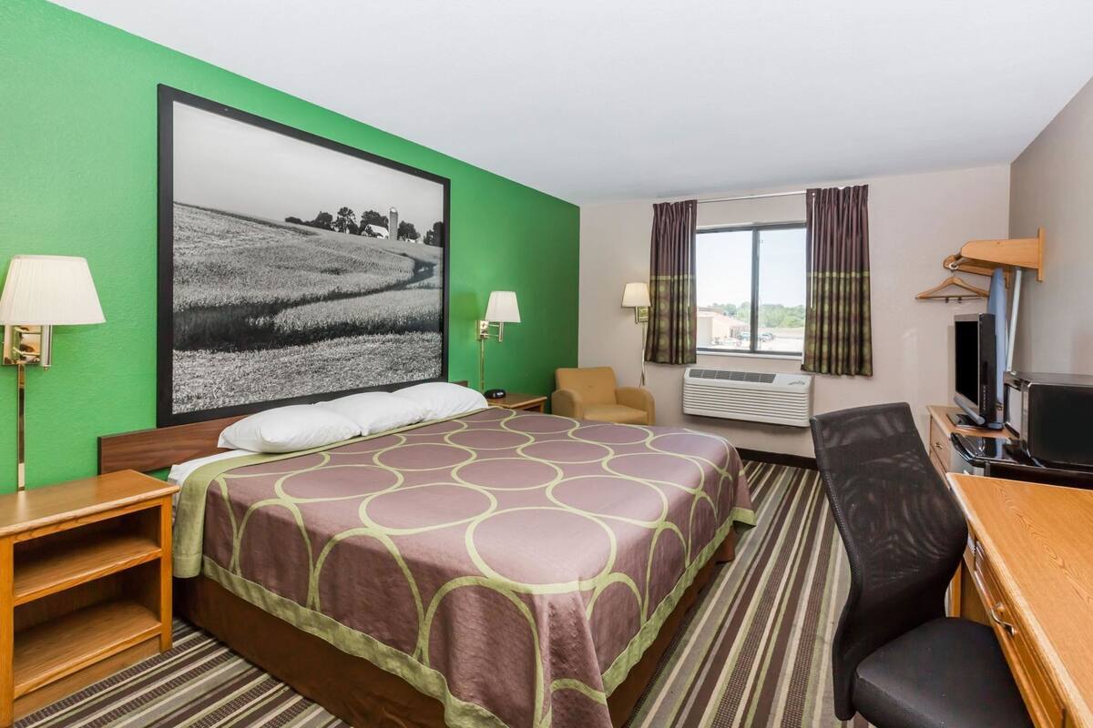 Habitación de hotel con cama, escritorio, silla, televisión y un amplio ventanal con cortinas.