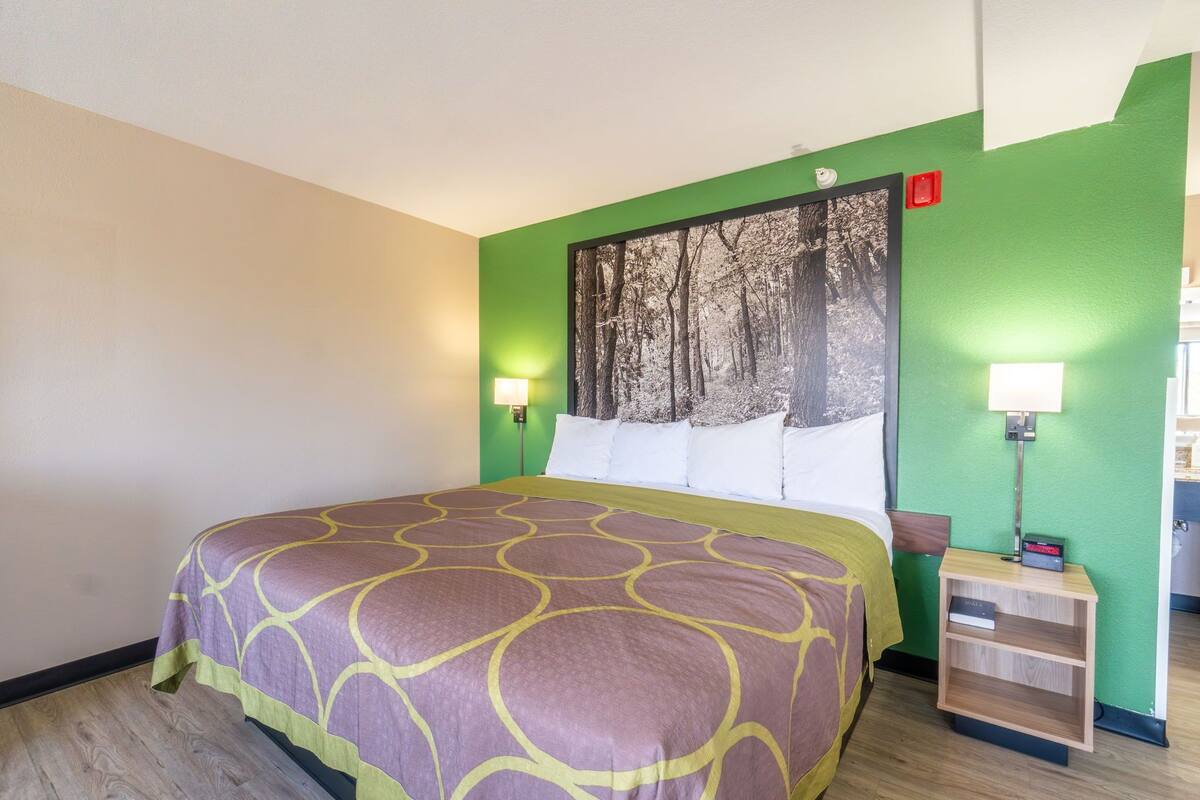 Habitación de hotel con cama, paredes verdes, cabecera con motivos de bosque, dos lámparas de noche y una mesita de noche con un libro.
