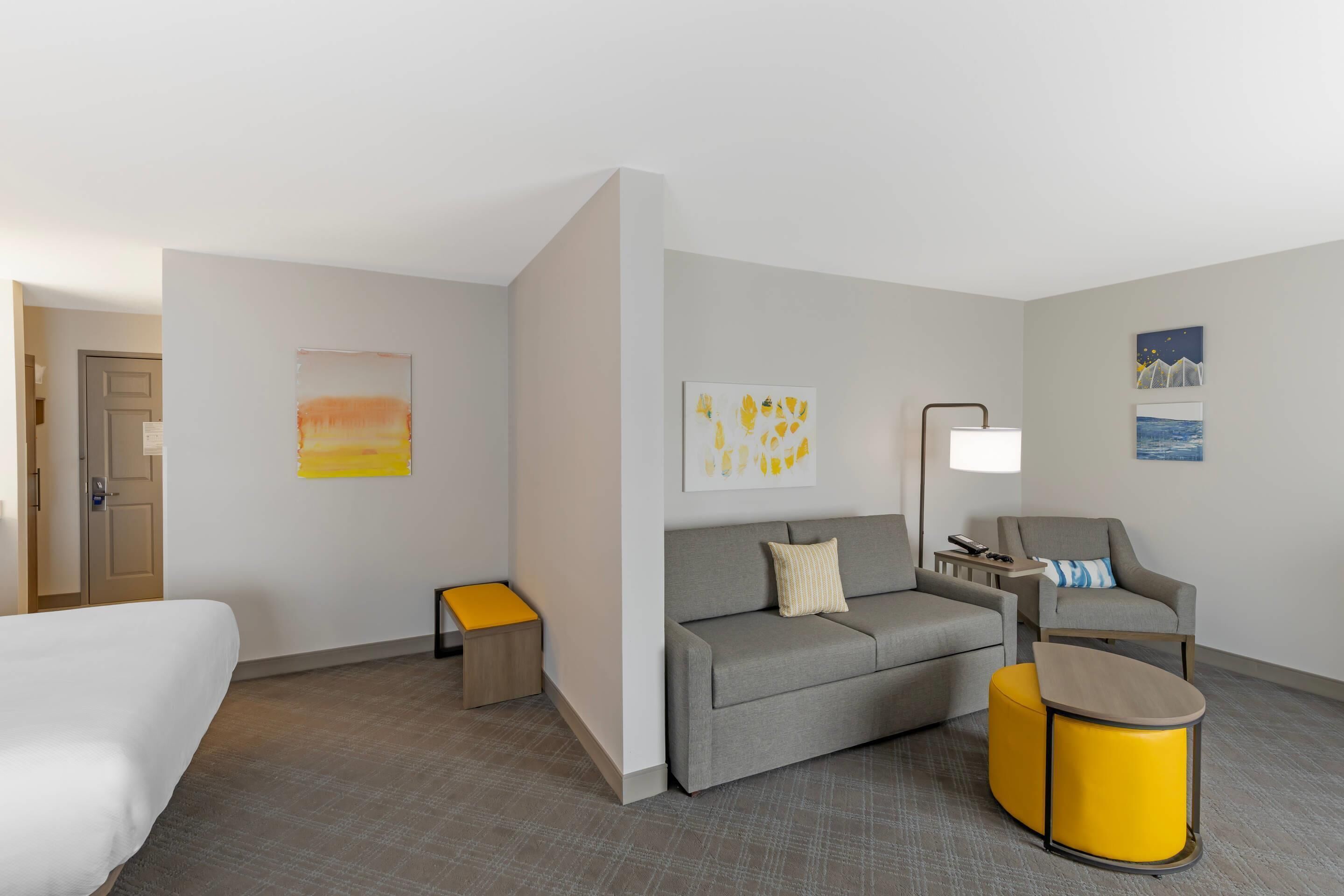 Quarto de hotel com cama, sofá, poltrona, mesa pequena e obras de arte nas paredes.