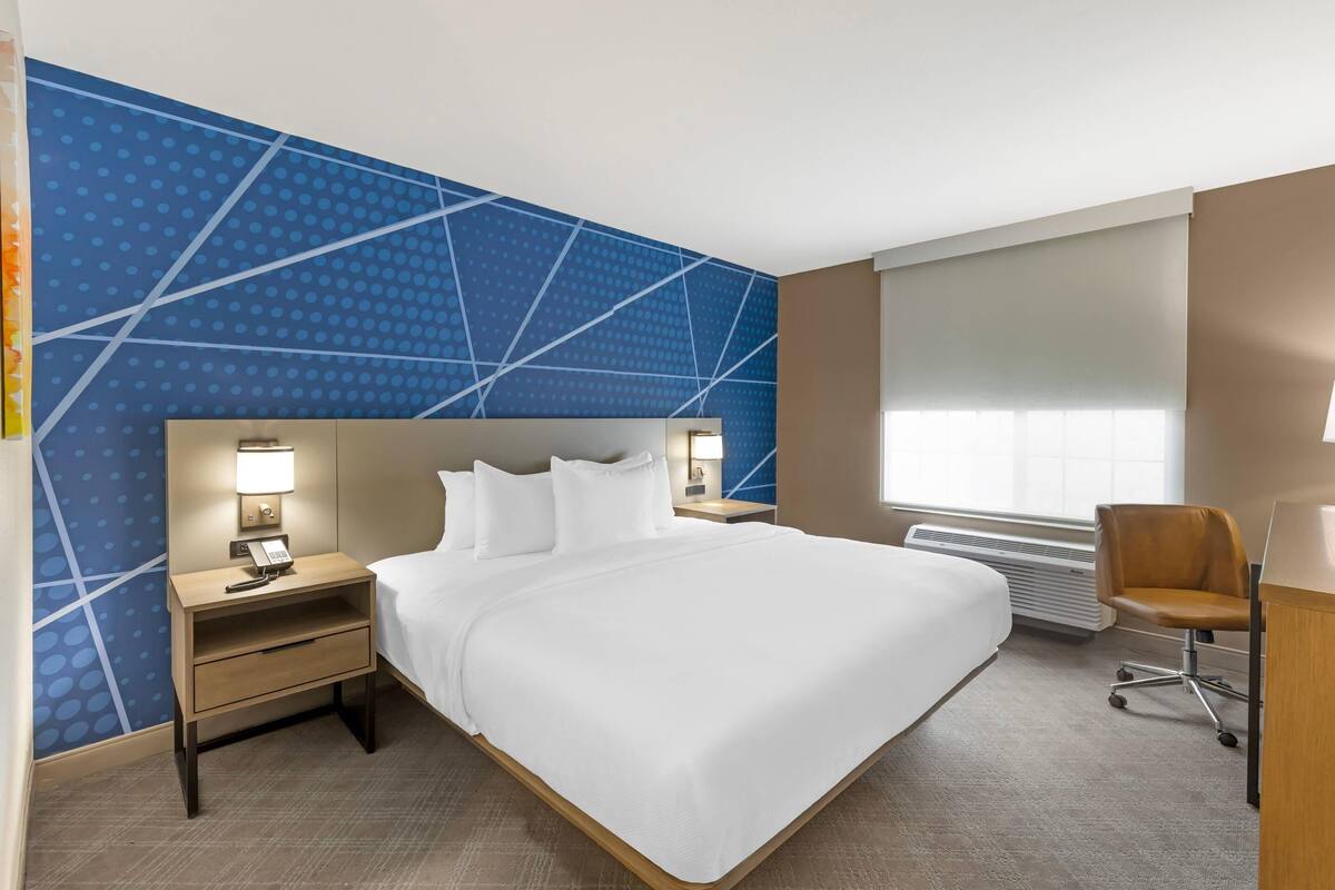 Une chambre d’hôtel avec un grand lit, un bureau, une chaise, une table de chevet avec une lampe et une fenêtre avec des stores.