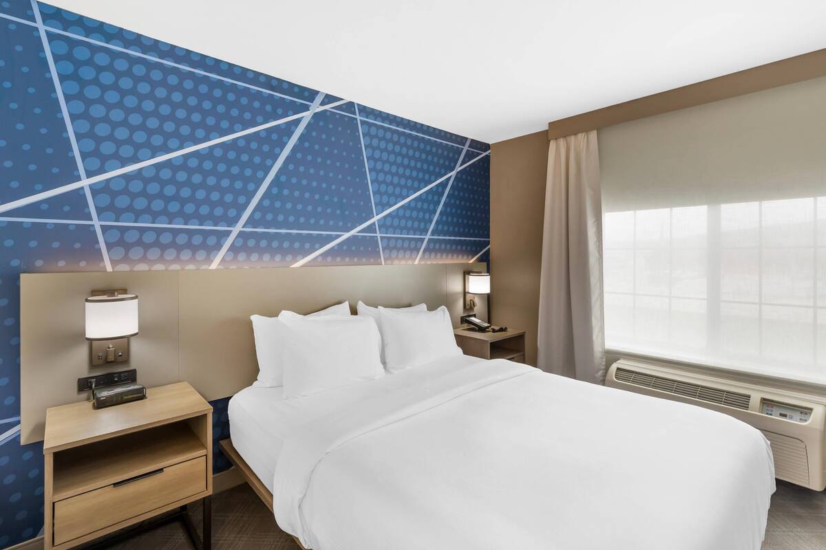 Une chambre d’hôtel avec un grand lit, une table de chevet avec une lampe, un climatiseur fixé au mur et une fenêtre avec des rideaux.