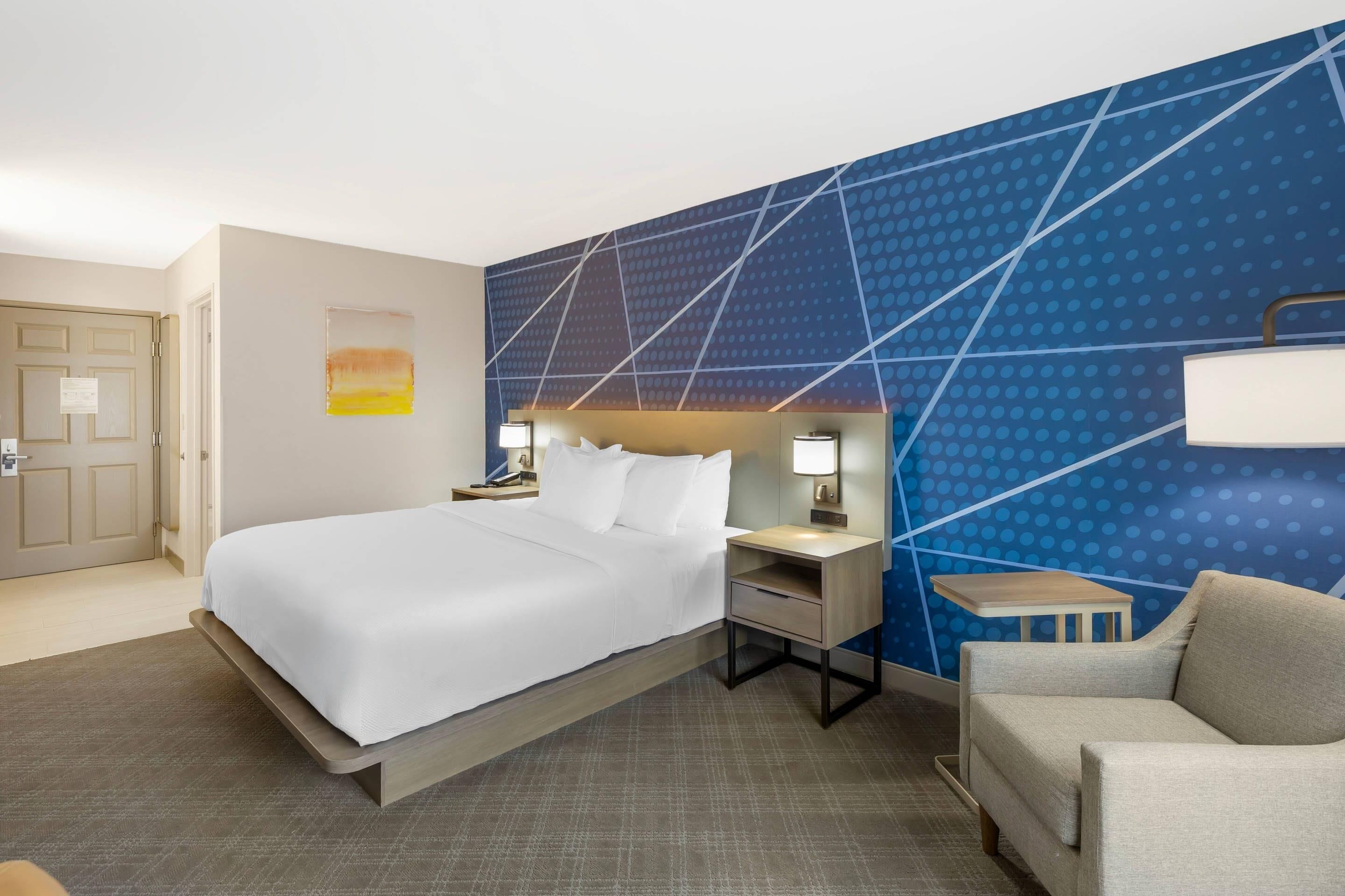 A hotel room with a bed, bedside table, lamp, chair, and a wall with a geometric pattern.