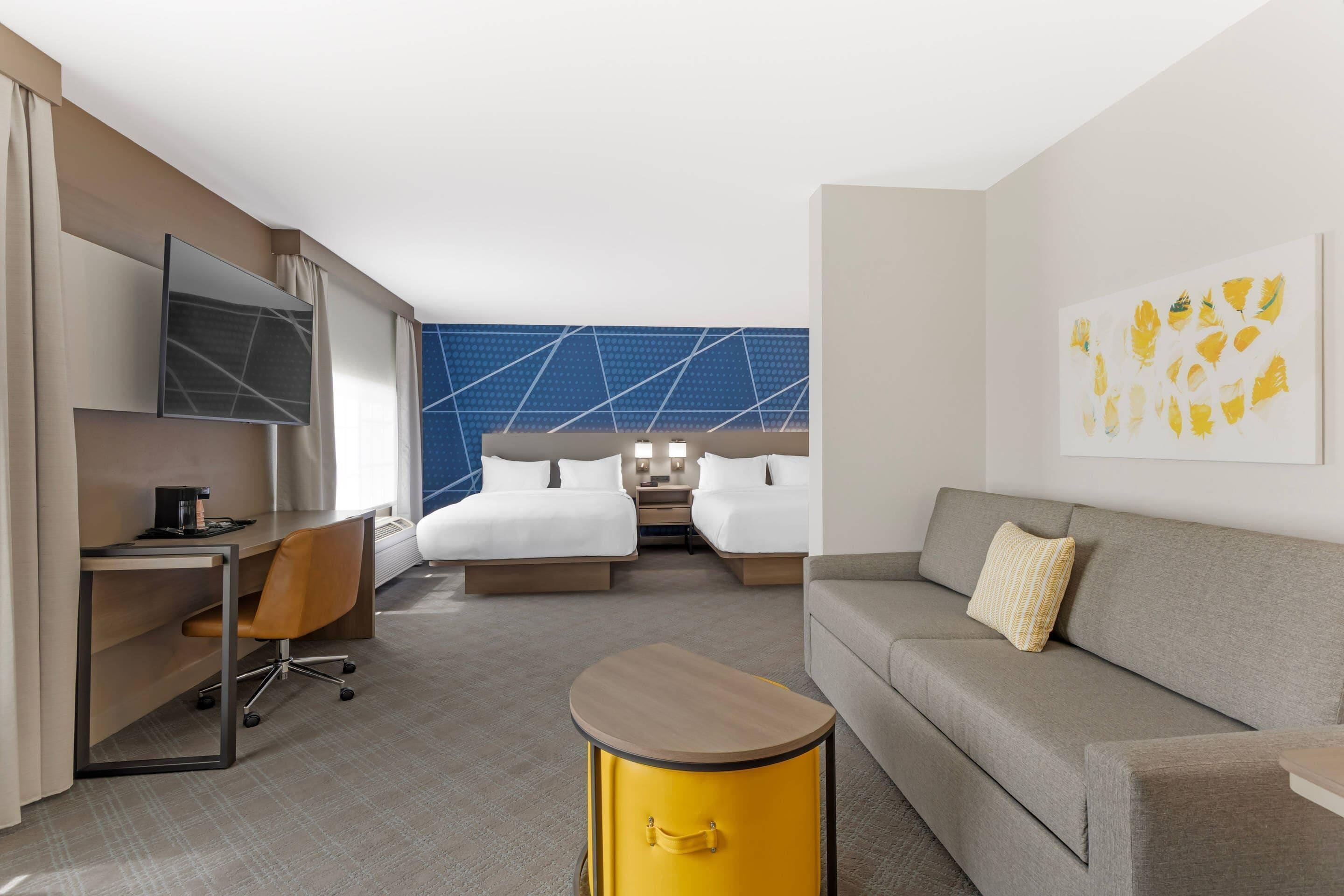 Habitación de hotel con cama, sofá, escritorio, silla, mesita, televisor y una pared con diseño geométrico.