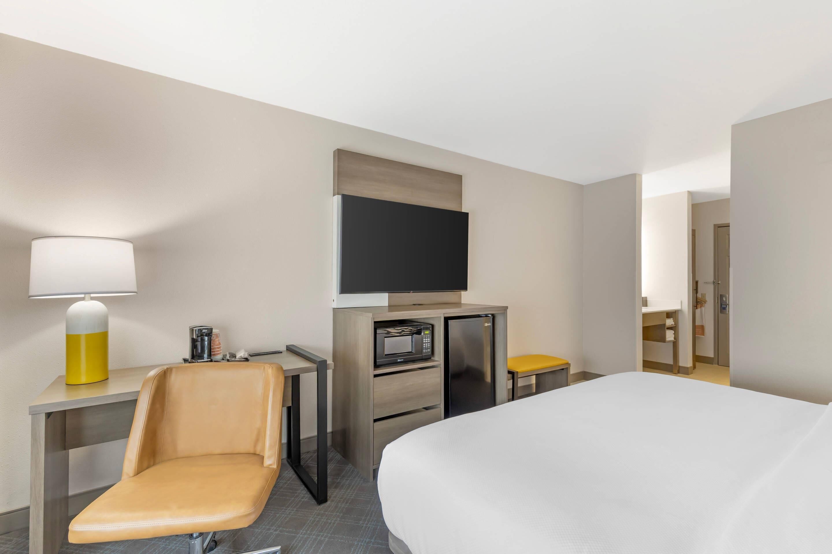 A hotel room with a bed, a desk with a lamp, a microwave, a TV, and a chair.