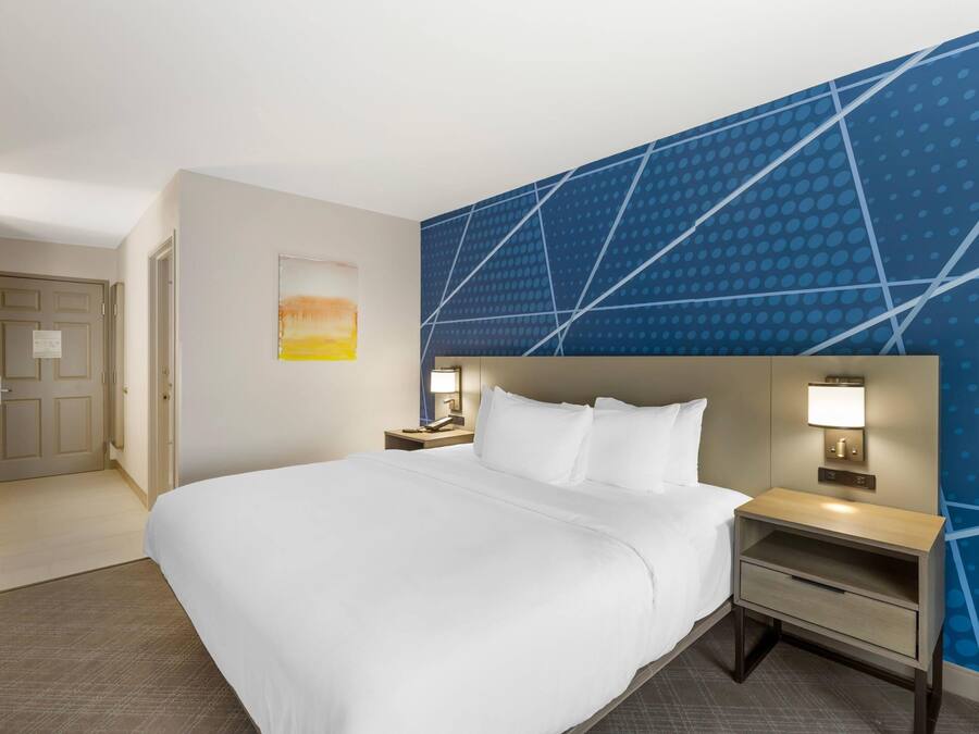 Ein Hotelzimmer mit einem großen Bett, einem Nachttisch mit Lampe, einem an der Wand befestigten Fernseher und einer Tür zu einem weiteren Raum.