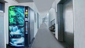 Un couloir avec un distributeur automatique affichant une image de cascade, un escalier et une porte sur la droite.