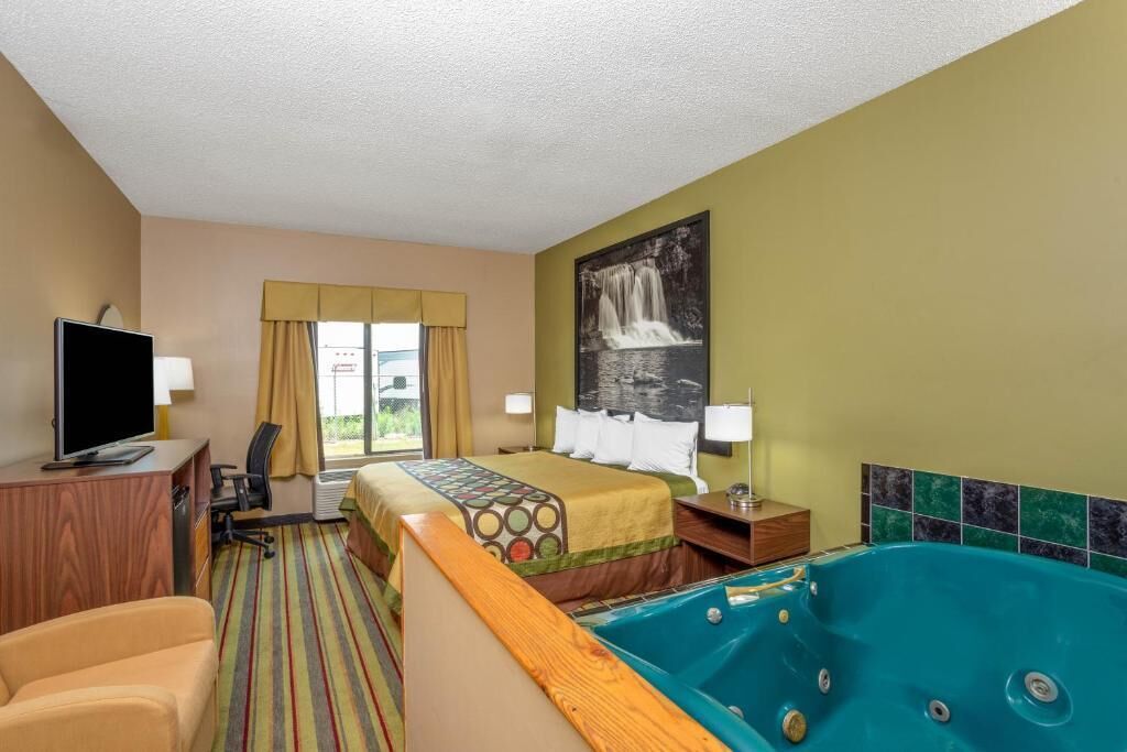 A hotel room with a bed, a desk with a computer, a chair, a large window with curtains, a painting of a waterfall, and a jacuzzi tub.