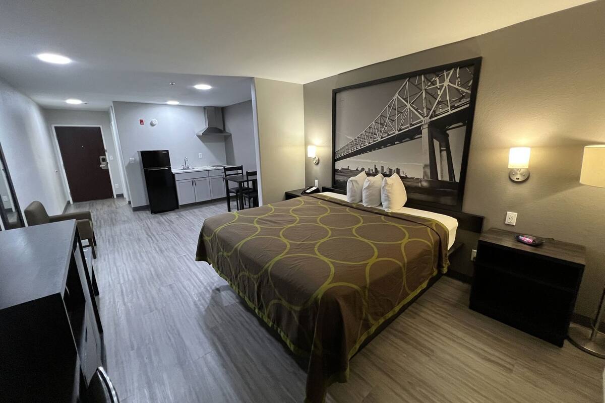 Habitación de hotel con cama, escritorio, silla, kitchenette pequeña y un amplio ventanal con vista a la ciudad.