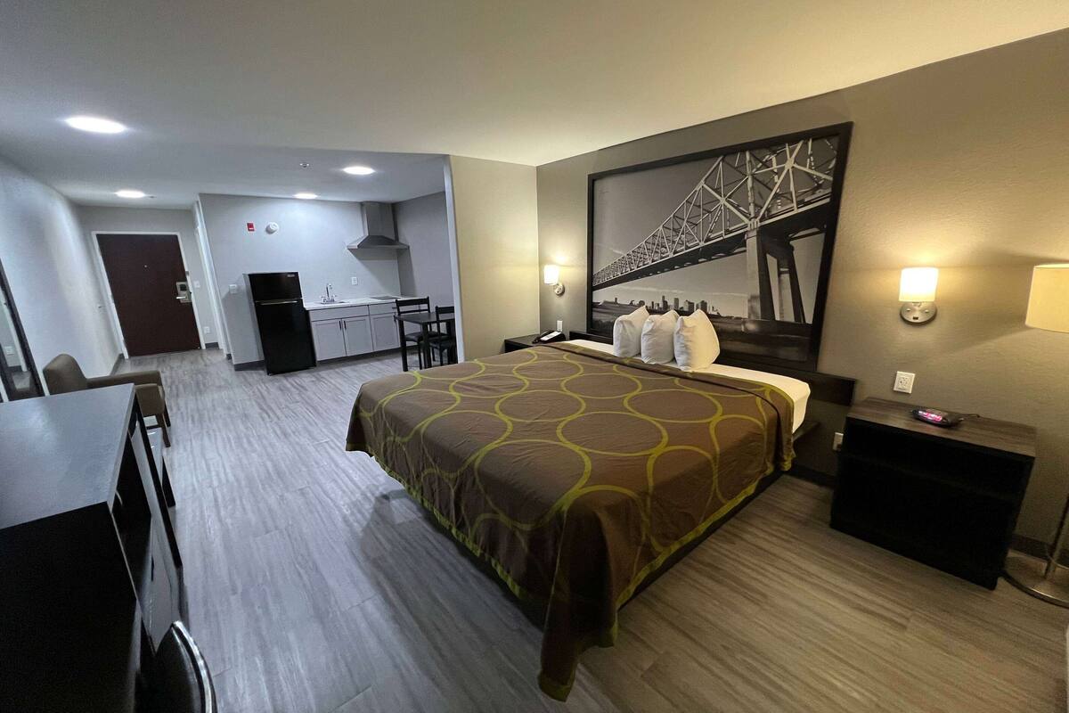 Habitación de hotel con una cama grande, un escritorio, una silla, una kitchenette pequeña y un ventanal con vistas a un puente.