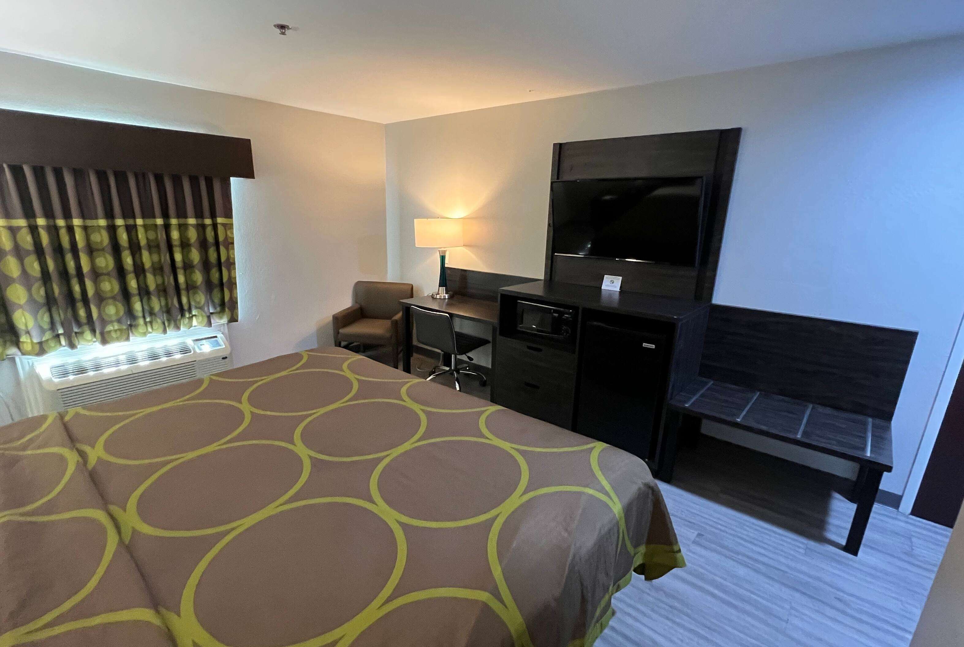 A hotel room with a large bed, a desk with a chair, a television, and a bench.