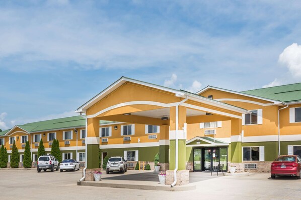 Un hotel de varios pisos con fachada amarilla y verde, una entrada cubierta y varios autos estacionados.
