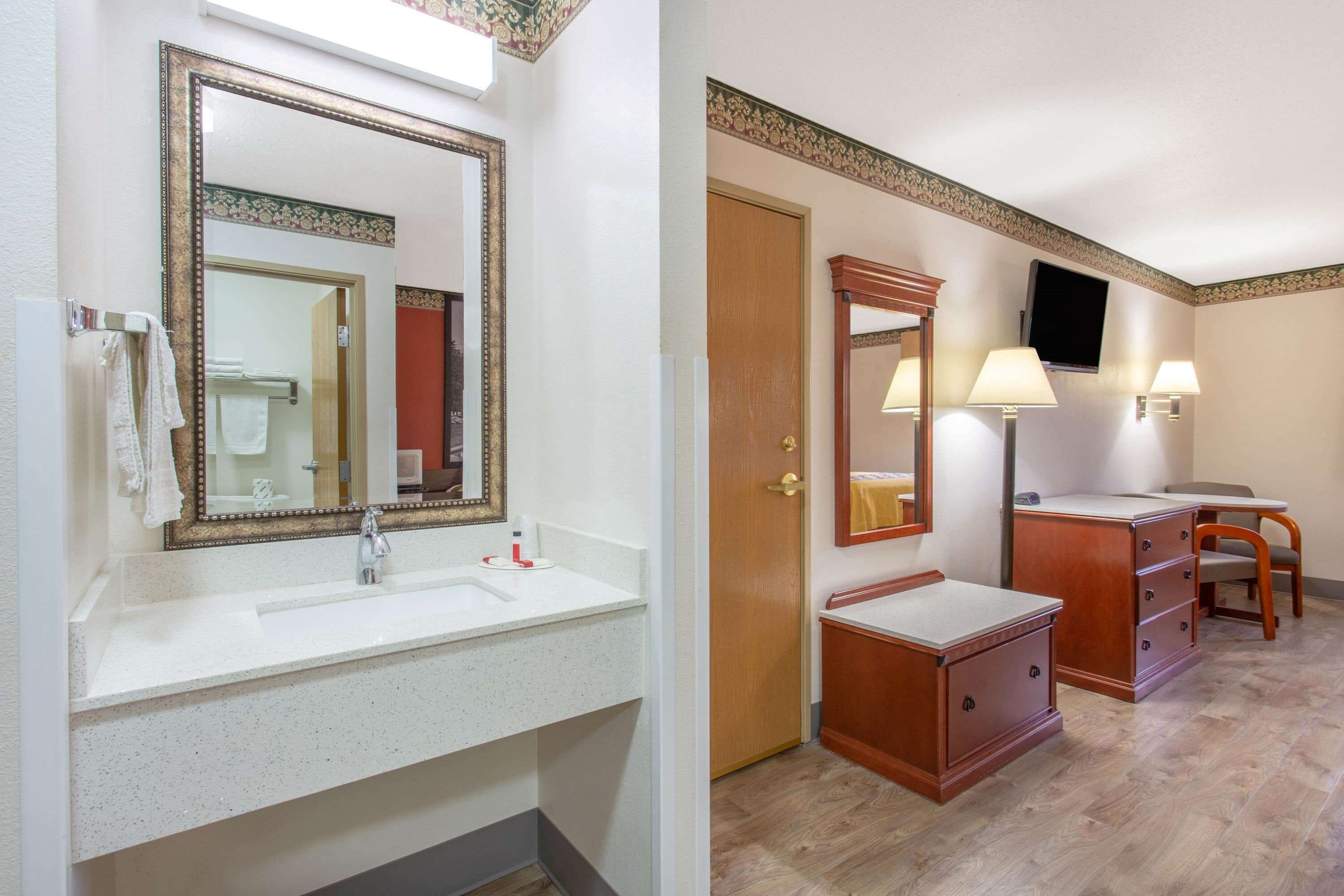 Un baño de hotel con un espejo grande, un lavamanos, un tocador con cajones, una silla y un televisor montado en la pared.