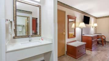 Un baño de hotel con un espejo grande, un lavamanos, un tocador con cajones, una silla y un televisor montado en la pared.