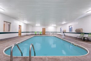 Una amplia piscina interior con agua cristalina y azul, rodeada de suelo y paredes con azulejos. Hay dos puertas a la izquierda, una ventana a la derecha y algunas sillas verdes cerca de la piscina.