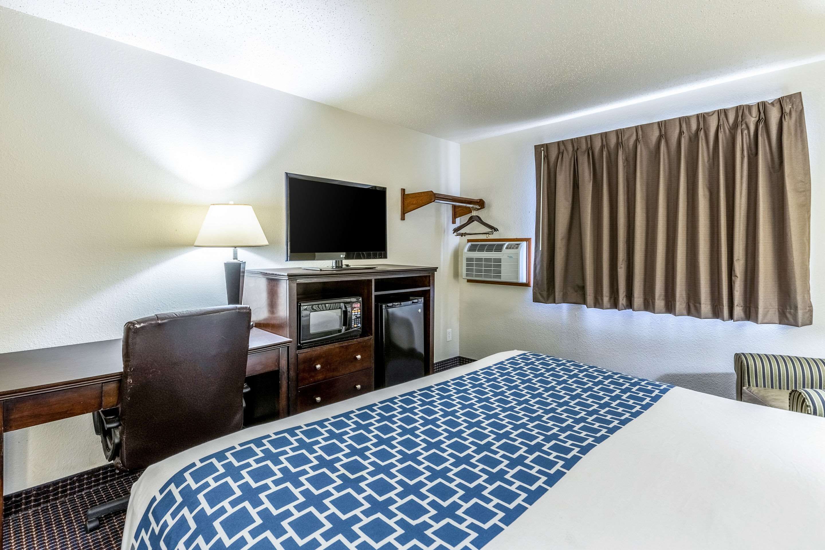 A hotel room with a bed, a desk with a computer, a television, a microwave, and an air conditioning unit.