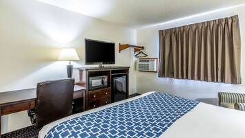 A hotel room with a bed, a desk with a computer, a television, a microwave, and an air conditioning unit.