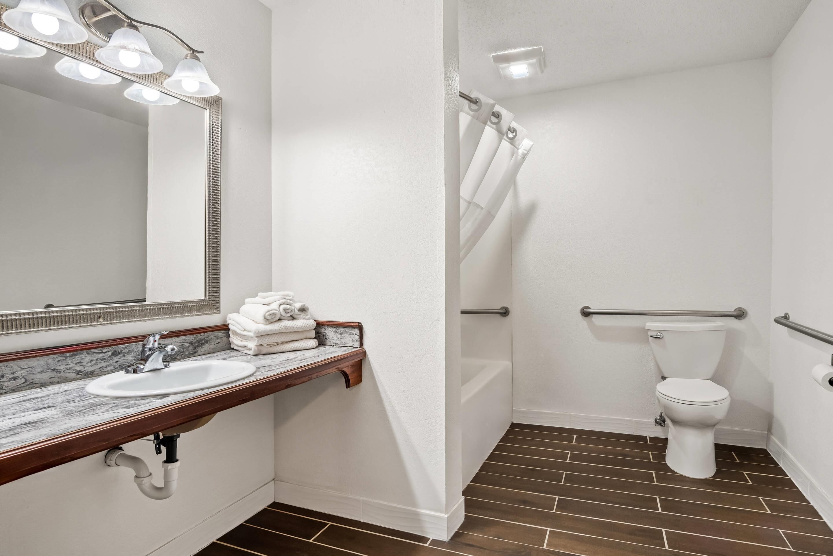 A bathroom with a white sink, mirror, and toilet. There are towels on the sink, a shower curtain, and a grab bar near the toilet.