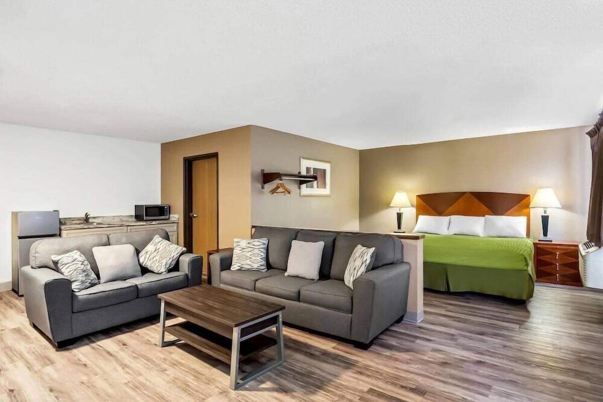 A modern hotel room with two sofas, a coffee table, a bed with a green bedspread, a microwave, a small kitchen area, and a wall-mounted TV.