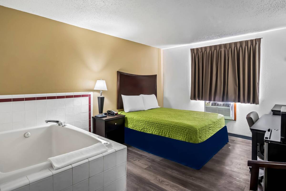 A hotel room with a large bathtub, a bed with a green and blue bedspread, a nightstand with a lamp, and a window with curtains.