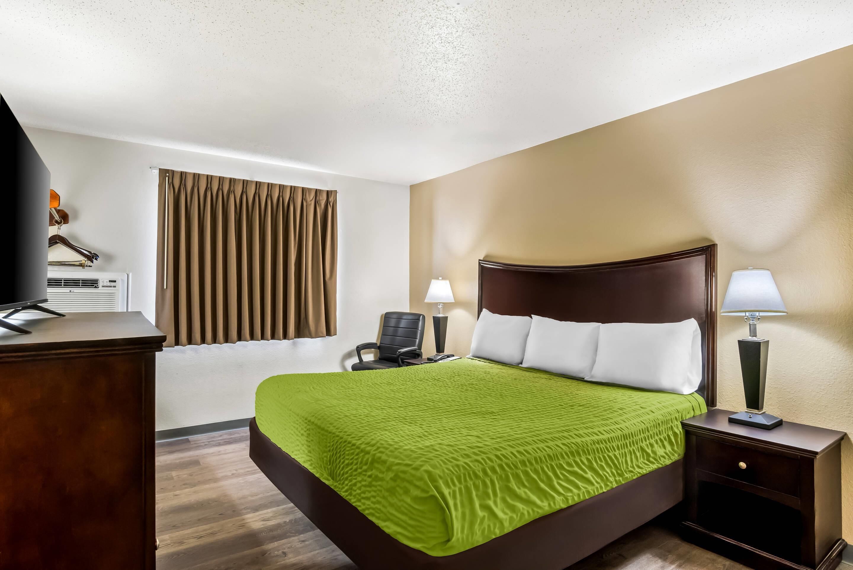 A hotel room with a bed, a desk with a television, a chair, a lamp, and a window with curtains.