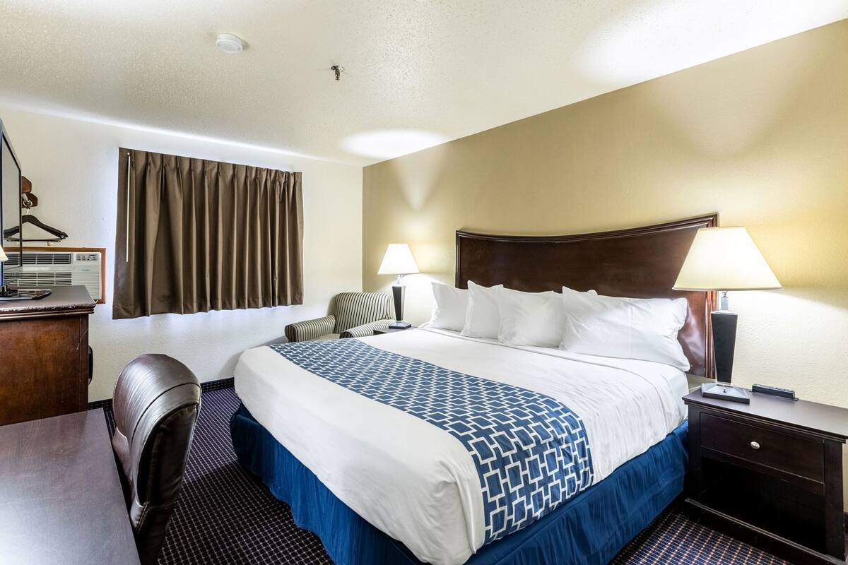 A hotel room with a large bed, two bedside lamps, a desk with a chair, a window with curtains, and a wall-mounted air conditioning unit.