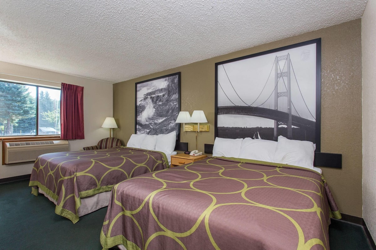 A hotel room with two beds, a large window with a view of trees, and two wall-mounted lamps.