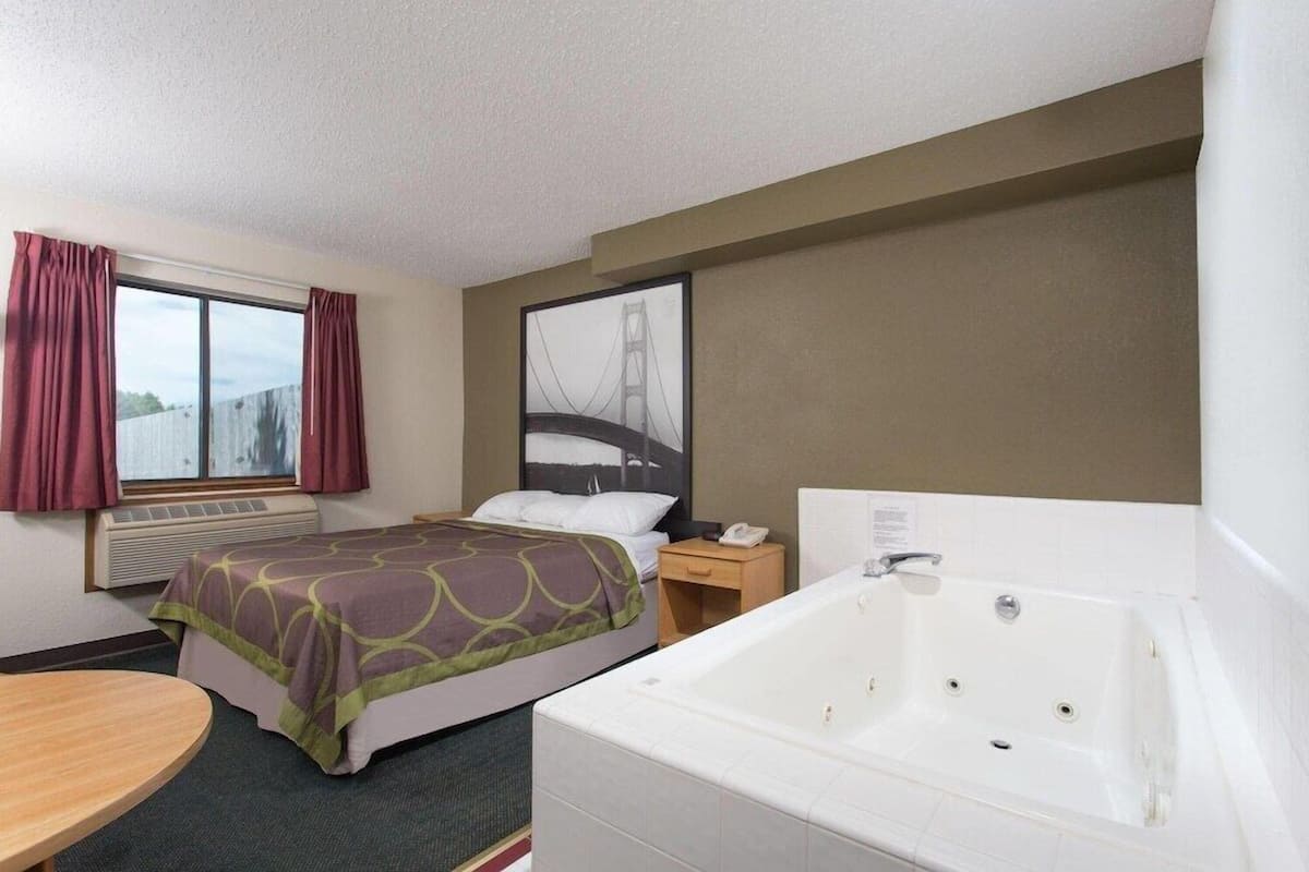 A hotel room with a bed, a bathroom with a bathtub, a window with curtains, and a wall-mounted air conditioner.