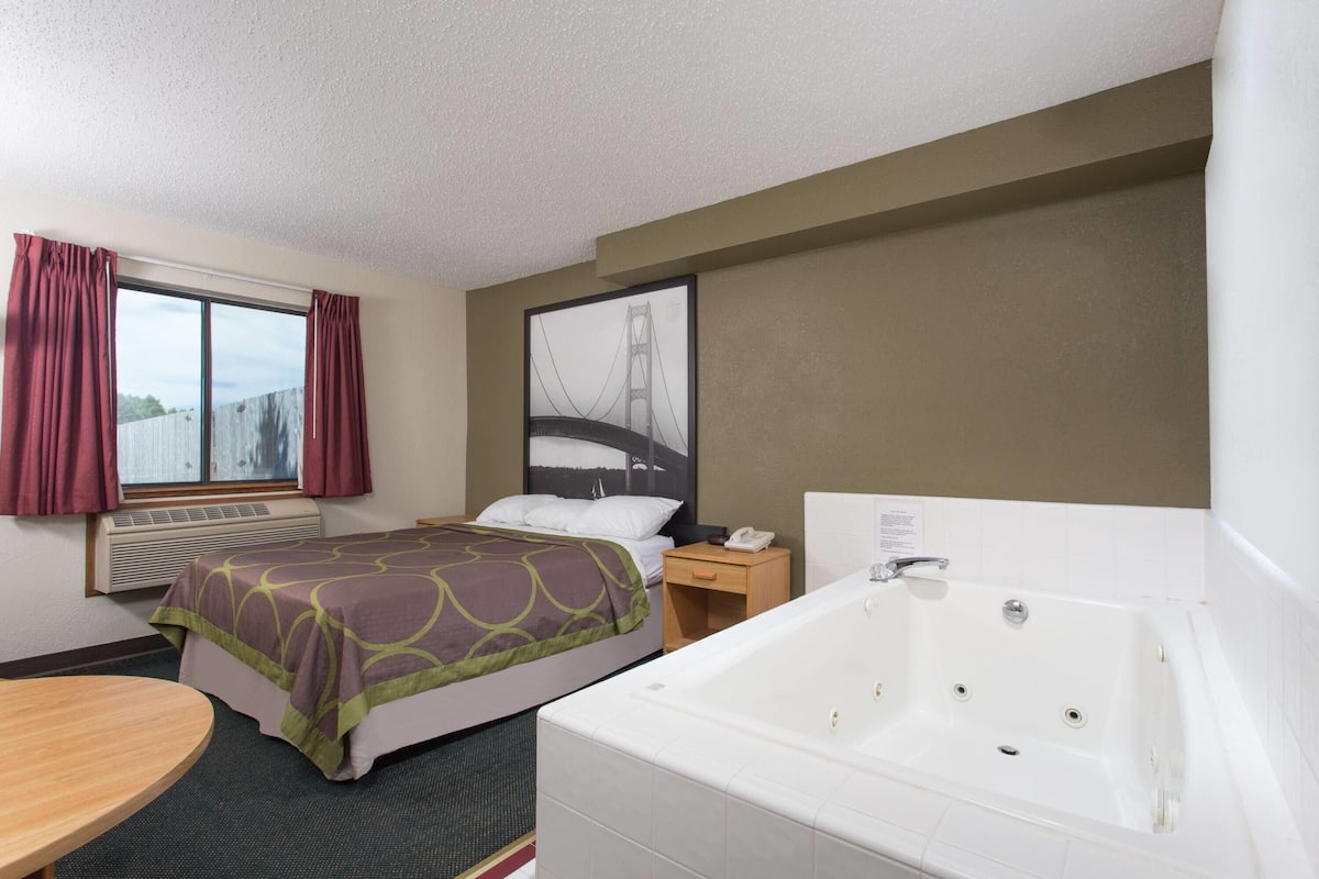 A hotel room with a bed, a large window with curtains, an air conditioning unit, a bedside table with a phone, and a bathtub.