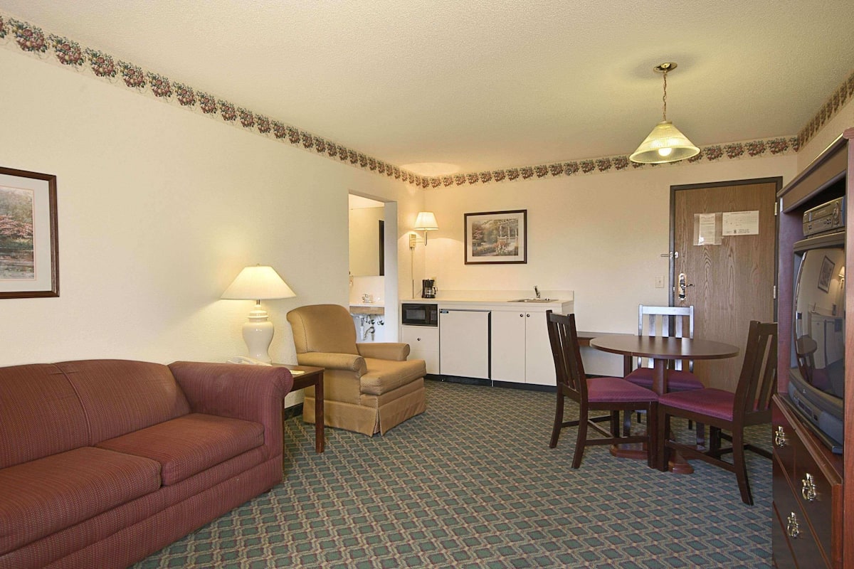 A compact hotel room with a sofa, a dining table with chairs, a kitchenette, and a television.