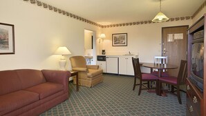 A compact hotel room with a sofa, a dining table with chairs, a kitchenette, and a television.