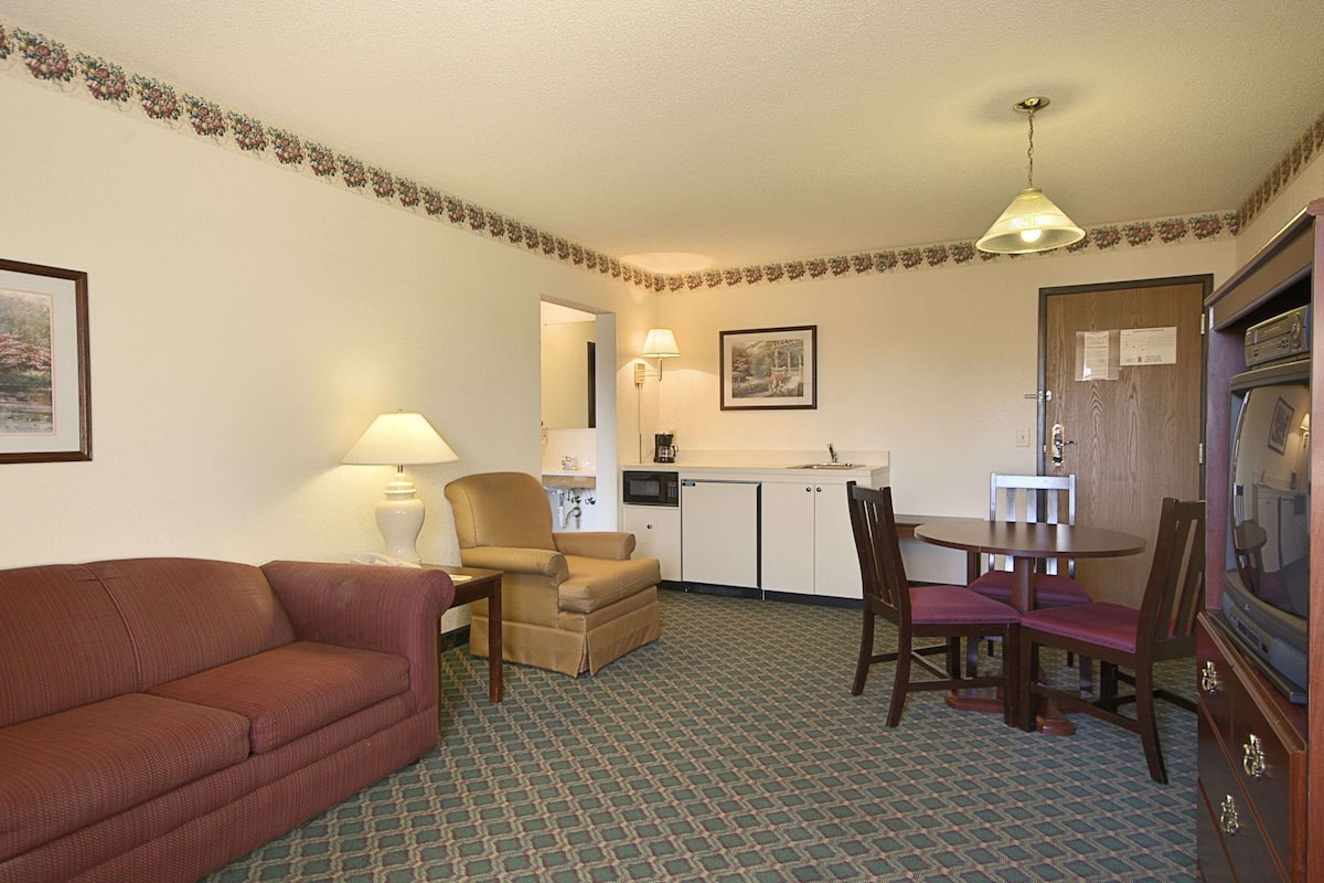 A compact hotel room with a sofa, a dining table with chairs, a kitchenette, and a television.