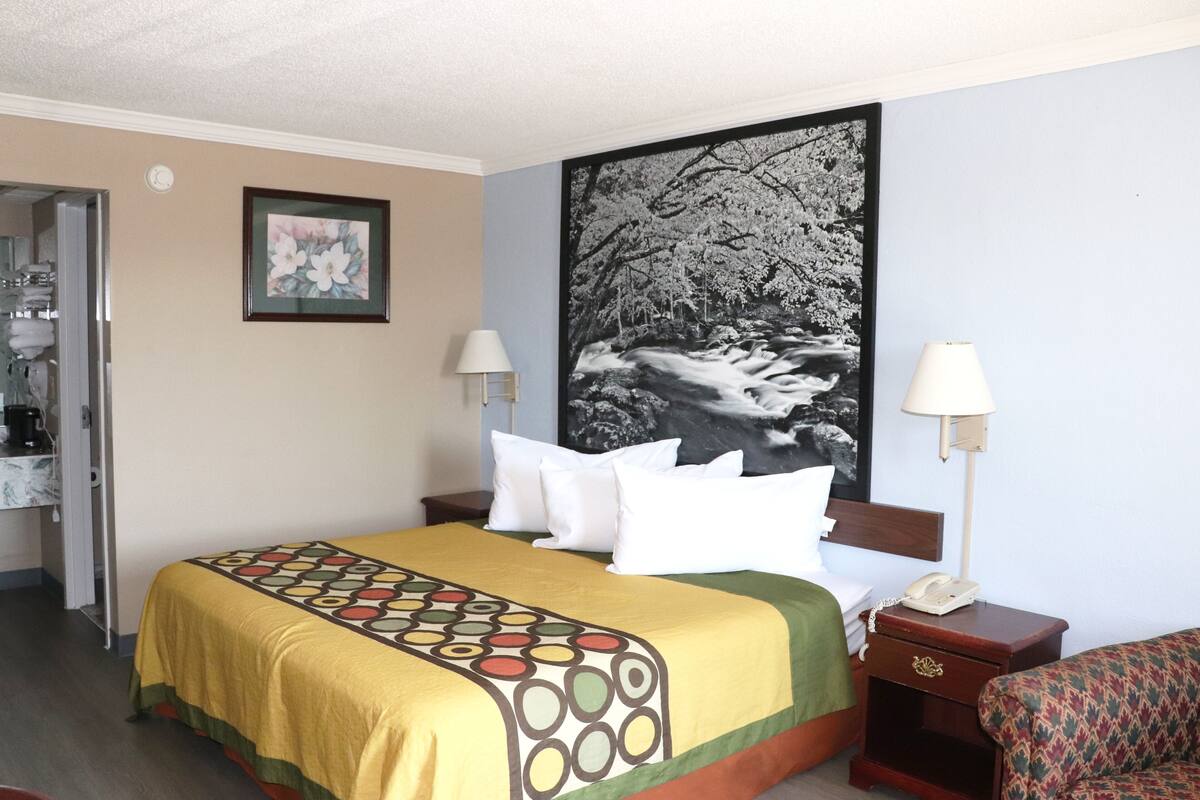 A hotel room with a bed, two bedside lamps, a nightstand with a phone, a framed picture on the wall, and a door leading to a bathroom.