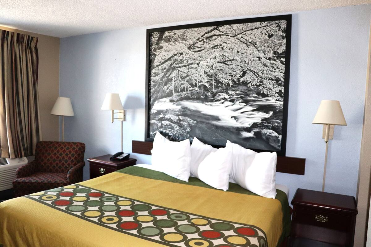 A hotel room with a bed, two bedside lamps, a chair, a nightstand, and a large framed picture on the wall.