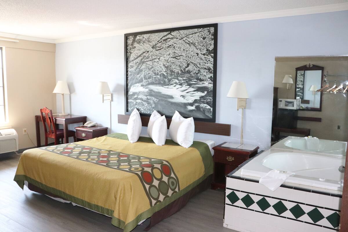 A hotel room with a bed, a desk, a chair, a desk lamp, a bedside table, a mirror, a bathroom with a bathtub, and a large framed picture on the wall.