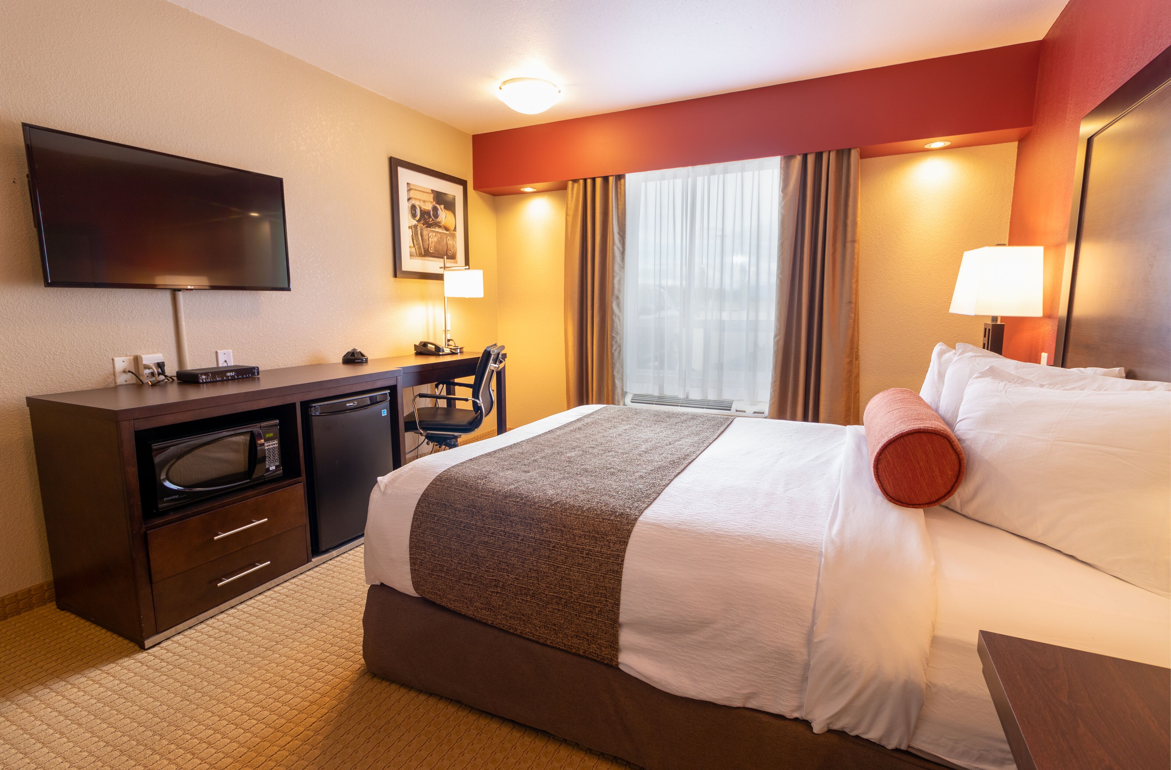 A hotel room with a bed, a desk with a microwave, a television, and a window with curtains.