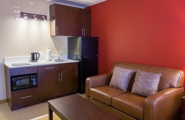 Cocina compacta con microondas, hervidor y refrigerador, junto a un sofá de cuero marrón con dos almohadas.