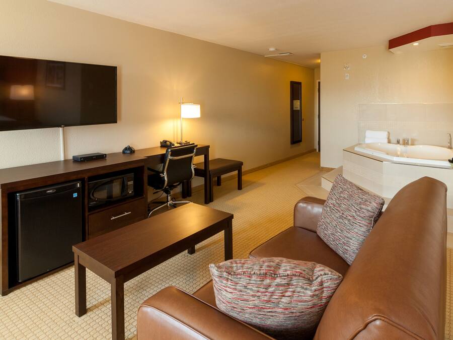 A hotel room with a flat-screen TV, a desk with a lamp, a sofa with a patterned pillow, a small refrigerator, and a bathtub with a towel.