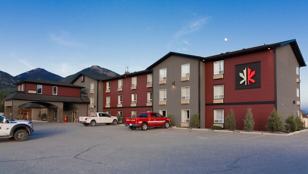 A multi-story hotel with a red and grey facade, a Canadian flag, and a parking lot with two trucks. - The Kanata by BCMInns Invermere (Invermere)