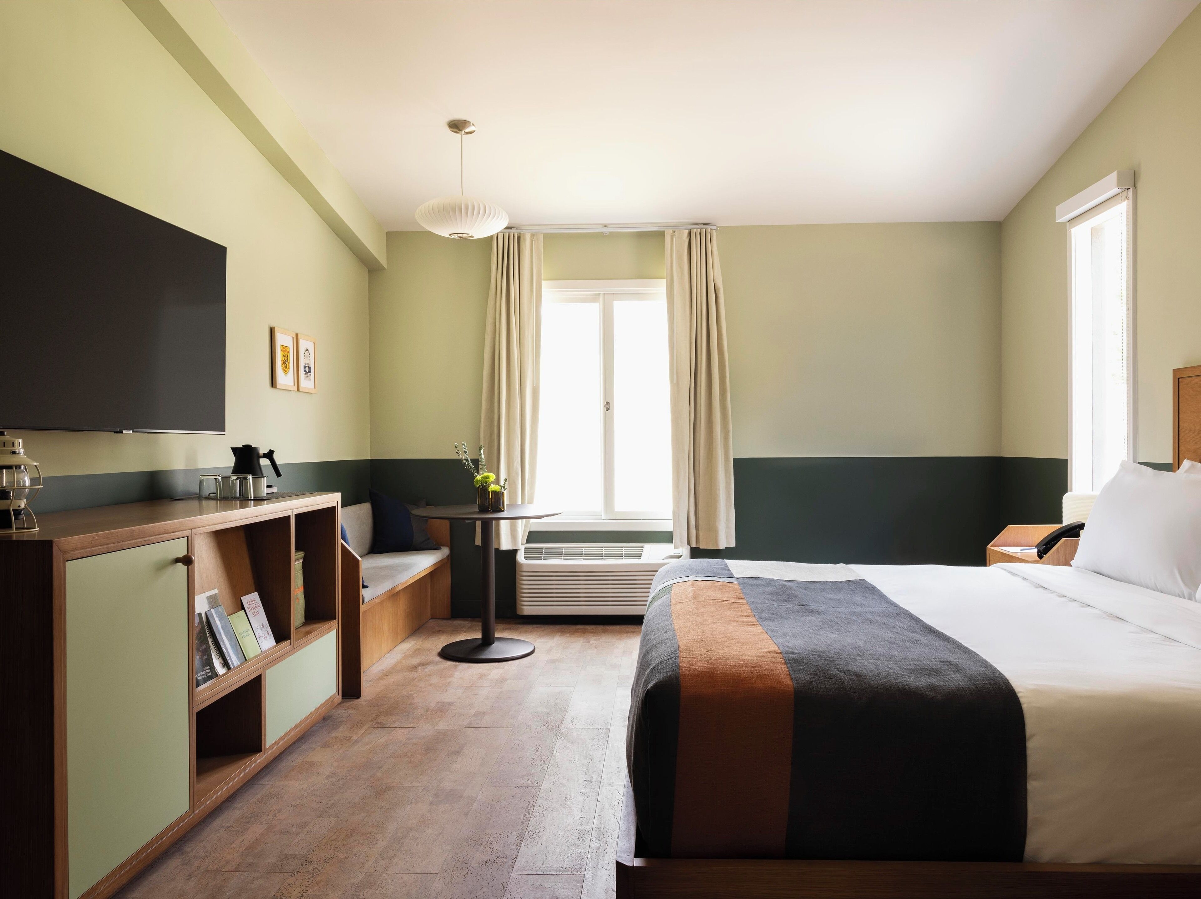 A hotel room with a bed, a desk with a chair, a bookshelf, a flat-screen TV, and a window with curtains.