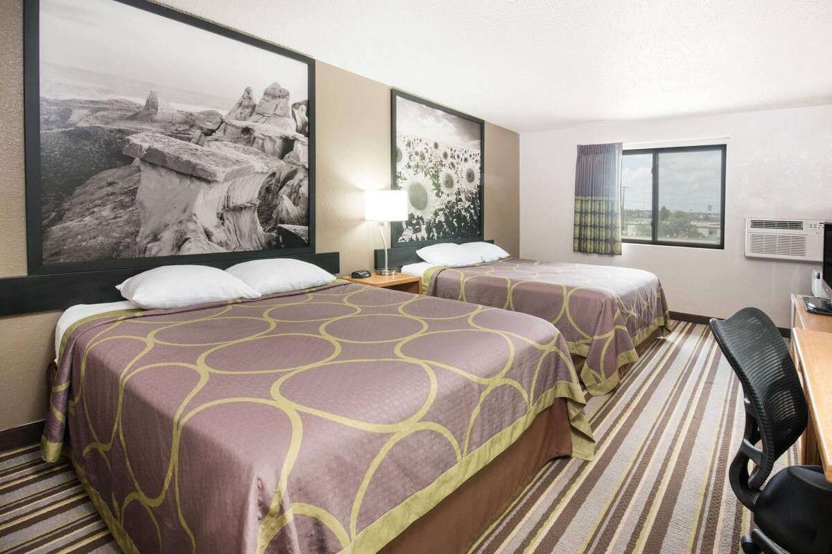 Une chambre d’hôtel avec deux lits, un bureau, une chaise, une lampe et un grand tableau encadré au mur.